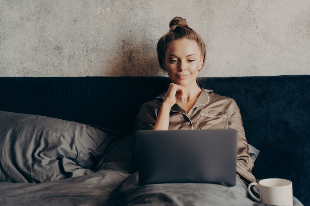Positive relaxed young woman in pajama lying in bed and using laptop